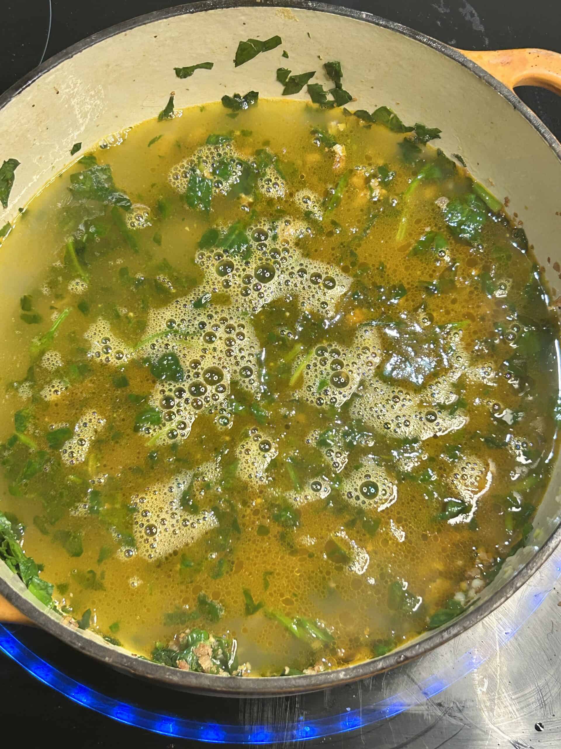 A dutch oven with broth and spinach, onion, ground sausage, and garlic