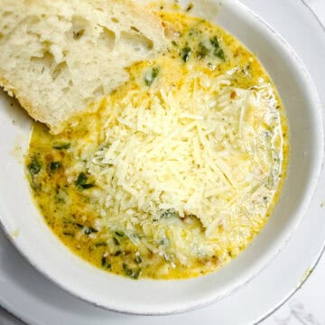 A bowl of creamy soup with visible herbs and greens, topped with shredded parmesan cheese. A slice of crusty bread with airy texture is partially dipped into the soup. The dish is served in a white bowl on a matching white plate, placed on a marble counter top