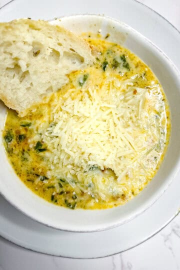 A bowl of creamy soup with visible herbs and greens, topped with shredded parmesan cheese. A slice of crusty bread with airy texture is partially dipped into the soup. The dish is served in a white bowl on a matching white plate, placed on a marble counter top