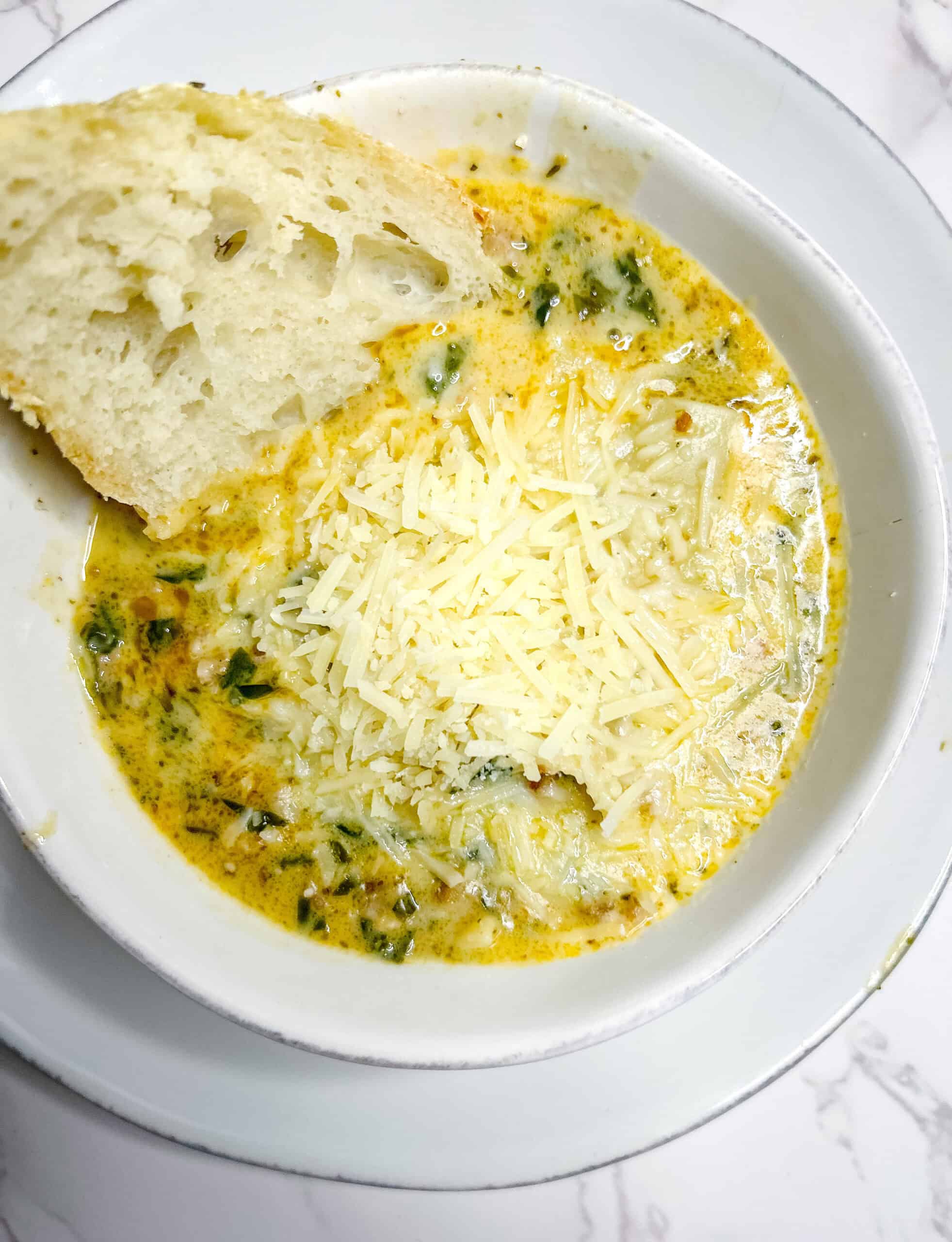 A bowl of creamy soup with visible herbs and greens, topped with shredded parmesan cheese. A slice of crusty bread with airy texture is partially dipped into the soup. The dish is served in a white bowl on a matching white plate, placed on a marble counter top