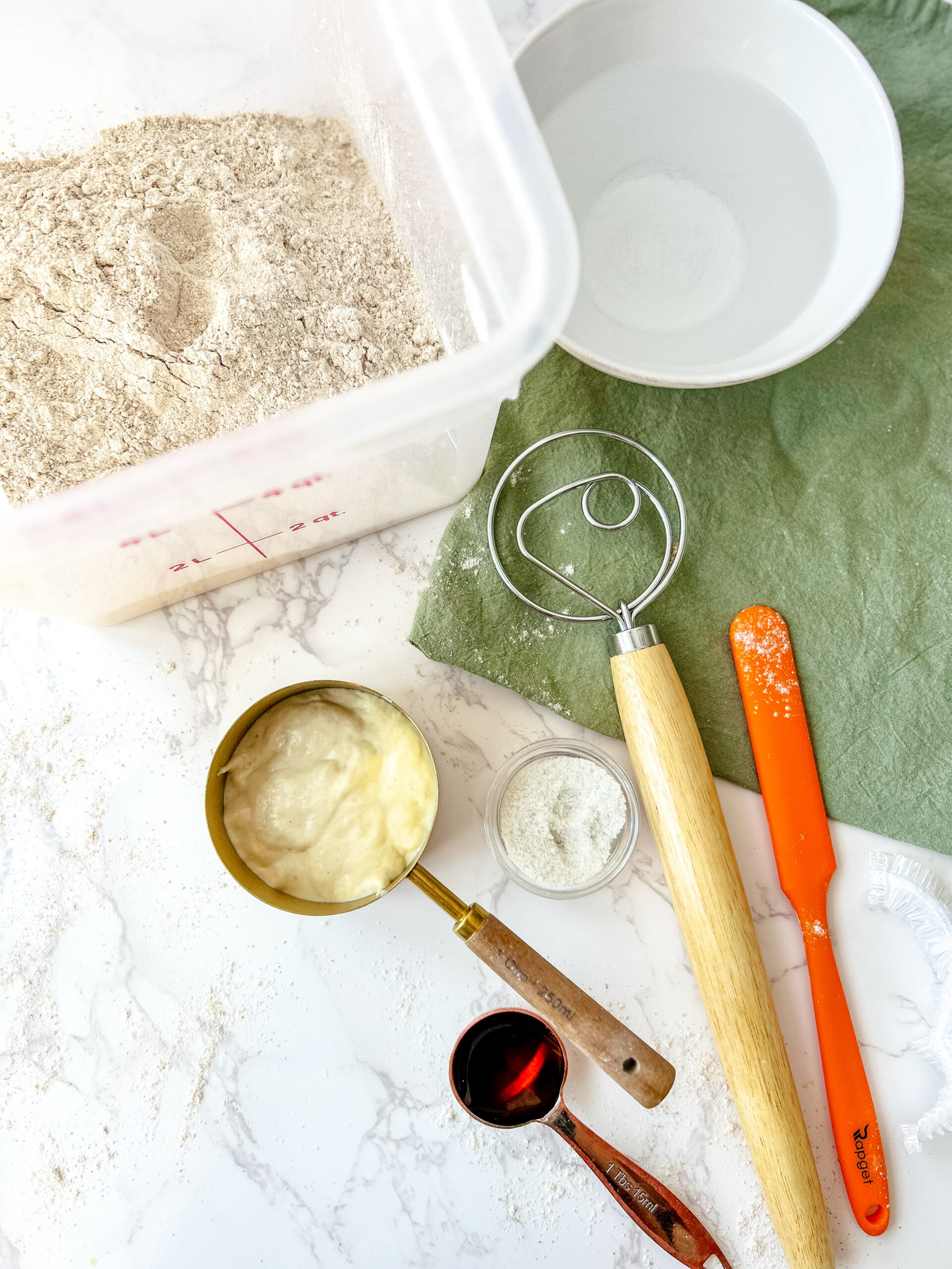 Ingredients and tools for making sourdough bread arranged on a white marble countertop. A container of freshly milled whole grain flour sits beside a bowl of water. A gold measuring cup holds sourdough starter, while small containers of salt and a dark liquid ingredient (possibly honey or molasses) are nearby. A Danish dough whisk, orange spatula, and wooden rolling pin rest on a green linen cloth, with scattered flour adding a rustic touch.
