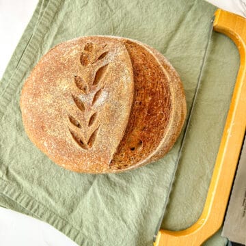 A round loaf of sourdough bread with a golden-brown crust, featuring a decorative wheat-scored pattern on top. The loaf is partially sliced, revealing a soft, airy interior. It rests on a muted green linen cloth atop a white marble surface. A serrated bread knife with a wooden handle is placed beside the bread.