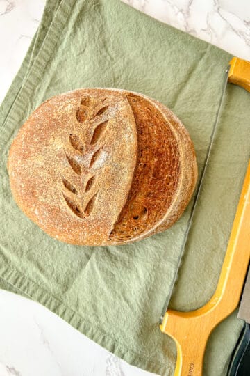 A round loaf of sourdough bread with a golden-brown crust, featuring a decorative wheat-scored pattern on top. The loaf is partially sliced, revealing a soft, airy interior. It rests on a muted green linen cloth atop a white marble surface. A serrated bread knife with a wooden handle is placed beside the bread.