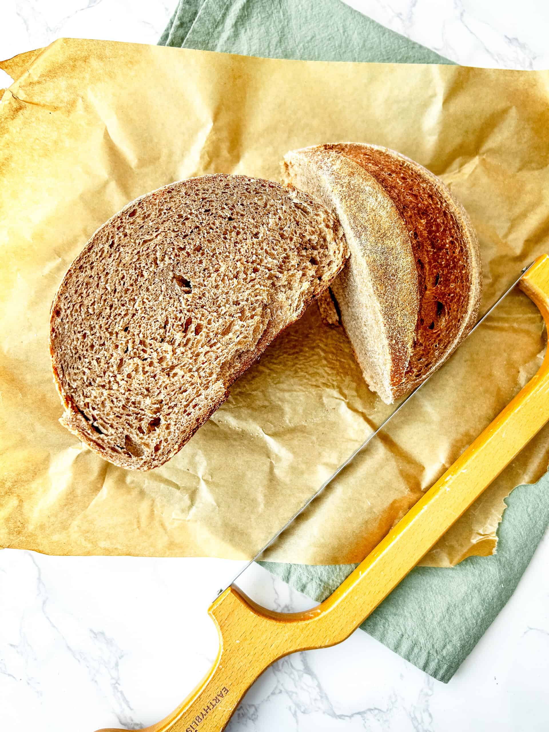 Sourdough bread that has been sliced in half on parchment paper. There is a rustic bread knife and a green tea towel.