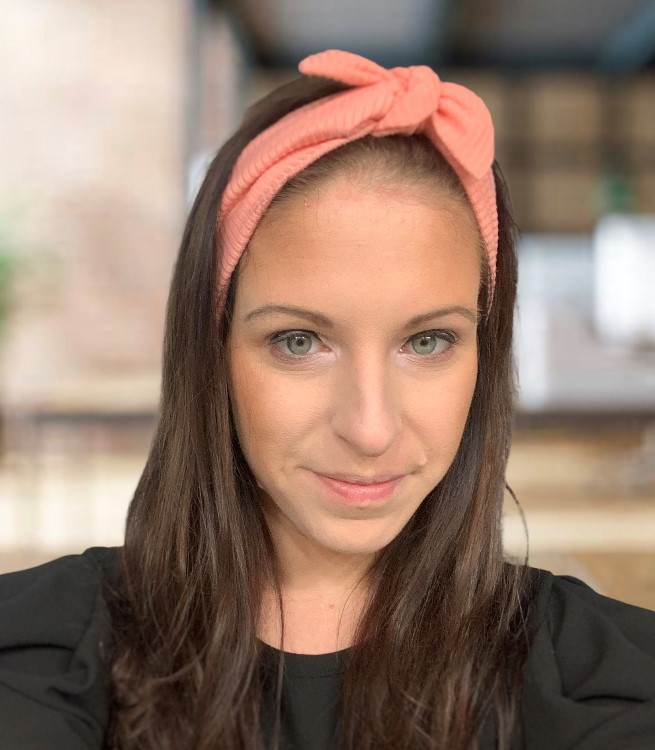 A woman with long brown hair wearing a peach-colored headband and a black top, smiling softly at the camera. The background is blurred, showing an indoor setting with warm lighting