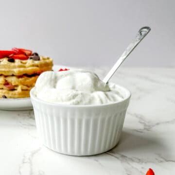 Whip cream in the front of the photo with a spoon sticking out, and a plate of waffles in the background with chocolate chips and strawberries on top