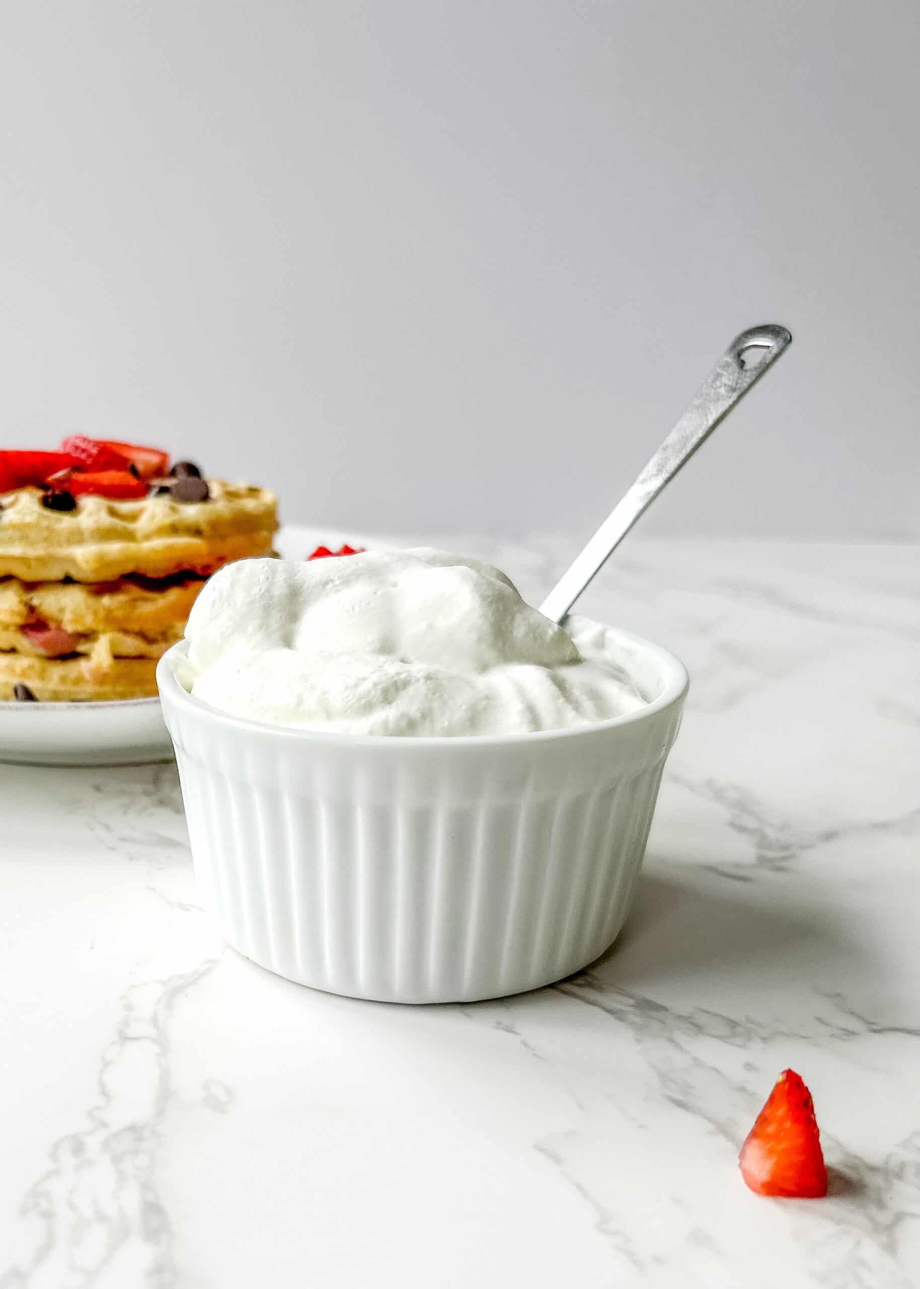 Whip cream in the front of the photo with a spoon sticking out, and a plate of waffles in the background with chocolate chips and strawberries on top